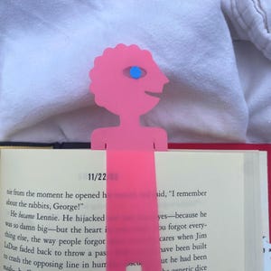 May include: A pink, whimsical bookmark featuring a stylized figure with a single blue eye. The bookmark is inserted into a book, with the date "11/22" visible. The bookmark's design adds a playful touch to reading.