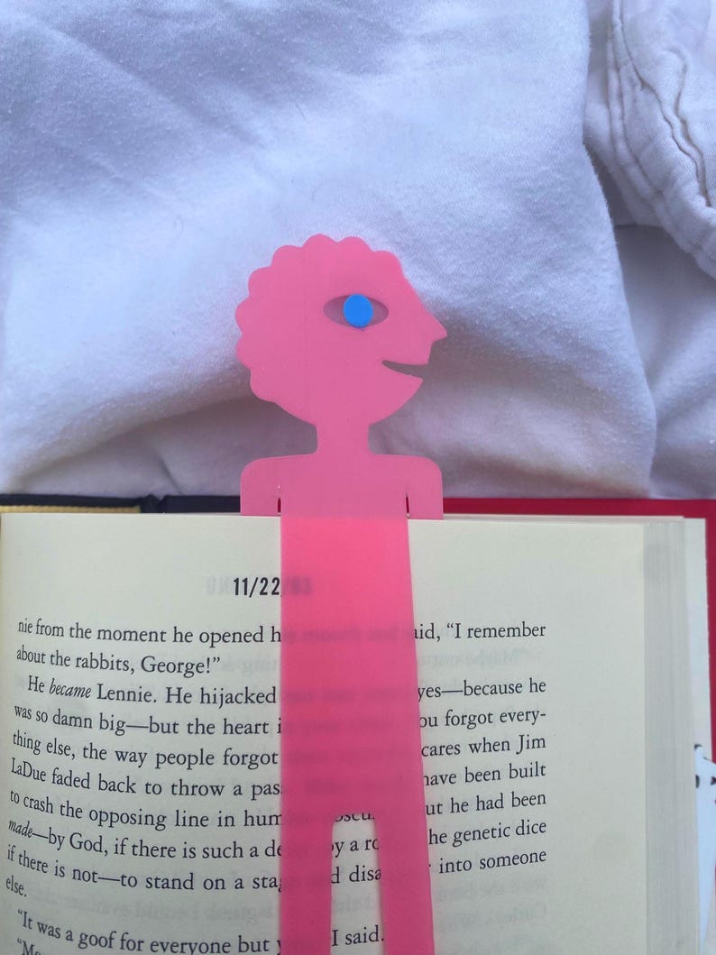 May include: A pink, whimsical bookmark featuring a stylized figure with a single blue eye. The bookmark is inserted into a book, with the date "11/22" visible. The bookmark's design adds a playful touch to reading.