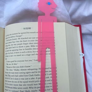 May include: A pink, stylized bookmark in the shape of a person, with a head, body, and legs, is inserted into a book. The bookmark has a blue eye and a smiling face. The book is open to a page with text.