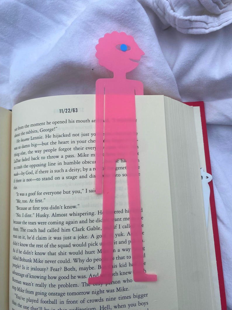 May include: A pink, stylized bookmark in the shape of a person, with a head, body, and legs, is inserted into a book. The bookmark has a blue eye and a smiling face. The book is open to a page with text.