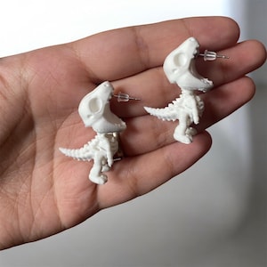 May include: White dinosaur skeleton earrings. Each earring features a small, detailed T-Rex skeleton design with a post for pierced ears. The earrings are a unique accessory, perfect for those who love dinosaurs or quirky jewelry.