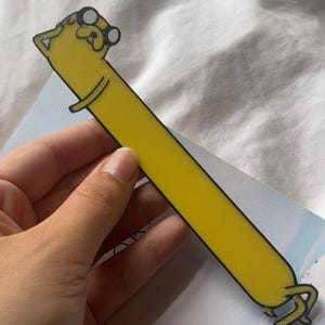 Peut inclure: Un marque-page jaune en forme du personnage de dessin anim&eacute; Jake le chien de la s&eacute;rie Adventure Time. Jake porte des lunettes et a un contour noir.