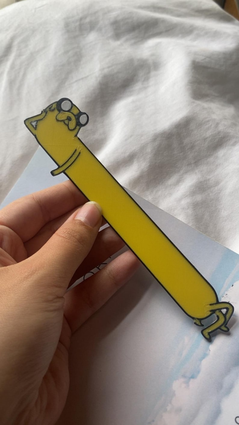Peut inclure: Un marque-page jaune en forme du personnage de dessin anim&eacute; Jake le chien de la s&eacute;rie Adventure Time. Jake porte des lunettes et a un contour noir.