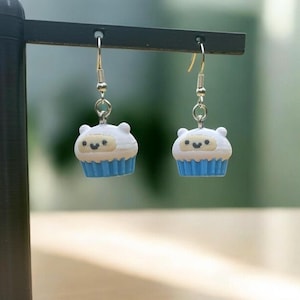 May include: A pair of novelty earrings featuring a cupcake design. The earrings have a white top with a smiling face and a blue cupcake liner. The earrings hang from gold-colored fish hook ear wires.