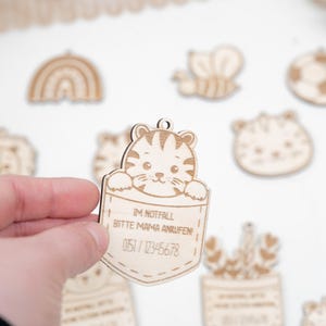 Könnte beinhalten: Holztiger-Taschenanhänger mit dem Text "Im Notfall Bitte Mama Anrufen 0151/12345678".
