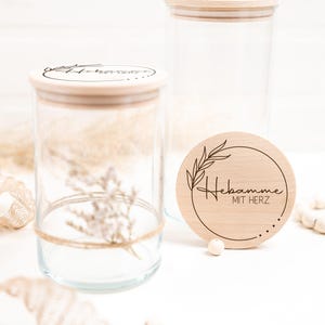 Könnte beinhalten: Zwei Glasbehälter mit Holzdeckeln. Der Deckel eines Behälters hat den Text "Hebamme mit Herz" eingraviert. Die Behälter stehen auf einer weißen Oberfläche mit einem Stück beigefarbenem Stoff und einer weißen Perlenkette.