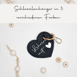 Könnte beinhalten: Schwarzer Leder-Schlüsselanhänger in Herzform mit goldenem Ring und Karabinerhaken. Der Schlüsselanhänger hat einen weißen Schriftzug "Lehrerin mit" und ein kleines weißes Herz. Der Schlüsselanhänger liegt auf einem weißen Hintergrund mit Holzperlen.