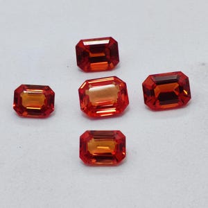 May include: Five rectangular-cut, orange-red gemstones are displayed against a white background. The gems have a vibrant, fiery colour with a polished, reflective surface. The facets catch the light, creating a sparkling effect. These gemstones are ideal for jewelry making.