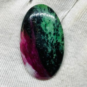 May include: An oval gemstone with a polished surface. The stone displays a mix of colours, including deep red, green, black, and white. The gemstone's pattern is a mix of swirling and speckled textures, creating a unique appearance. The stone is likely a natural mineral.