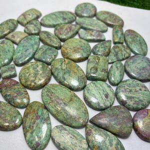Puede incluir: Una colección de piedras preciosas pulidas de varias formas, incluyendo óvalos, rectángulos y círculos. Las piedras tienen un color verde y rosa rojizo moteado, con un acabado liso y brillante. Las piedras preciosas son probablemente para la fabricación de joyas o coleccionar.