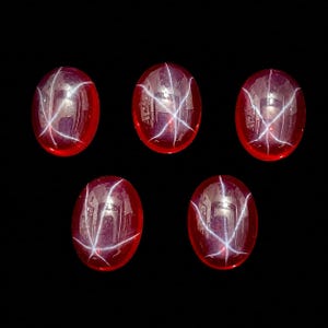 Peut inclure: Cinq pierres précieuses de forme ovale, taillées en cabochon, d'un rouge profond. Chaque gemme présente un effet d'étoile à six rayons, créé par des lignes de lumière qui se croisent. Les pierres sont posées sur un fond noir.