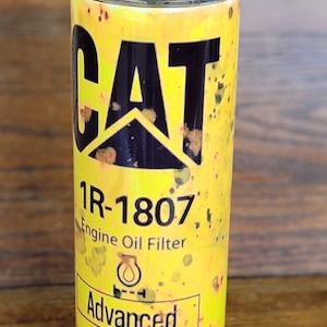 Może przedstawiać: Ż&oacute;łty kubek z czarnym logo "CAT" i napisem "1R-1807 Engine Oil Filter". Kubek ma przezroczystą pokrywkę i słomkę. Projekt zawiera słowo "Advanced" i "High Efficiency".