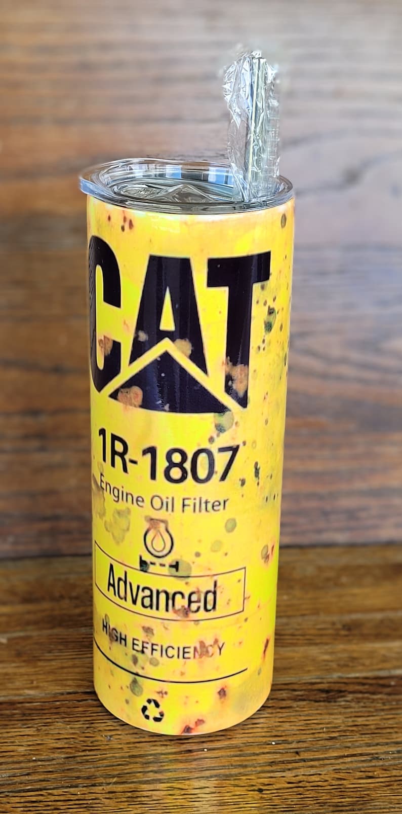 Może przedstawiać: Ż&oacute;łty kubek z czarnym logo "CAT" i napisem "1R-1807 Engine Oil Filter". Kubek ma przezroczystą pokrywkę i słomkę. Projekt zawiera słowo "Advanced" i "High Efficiency".