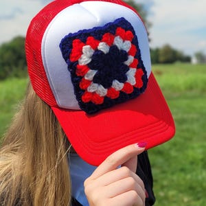 May include: A red and white trucker hat featuring a square crochet patch on the front panel. The patch is composed of concentric layers of dark blue, white, and red yarn in a classic granny square pattern, stitched onto the white foam front.