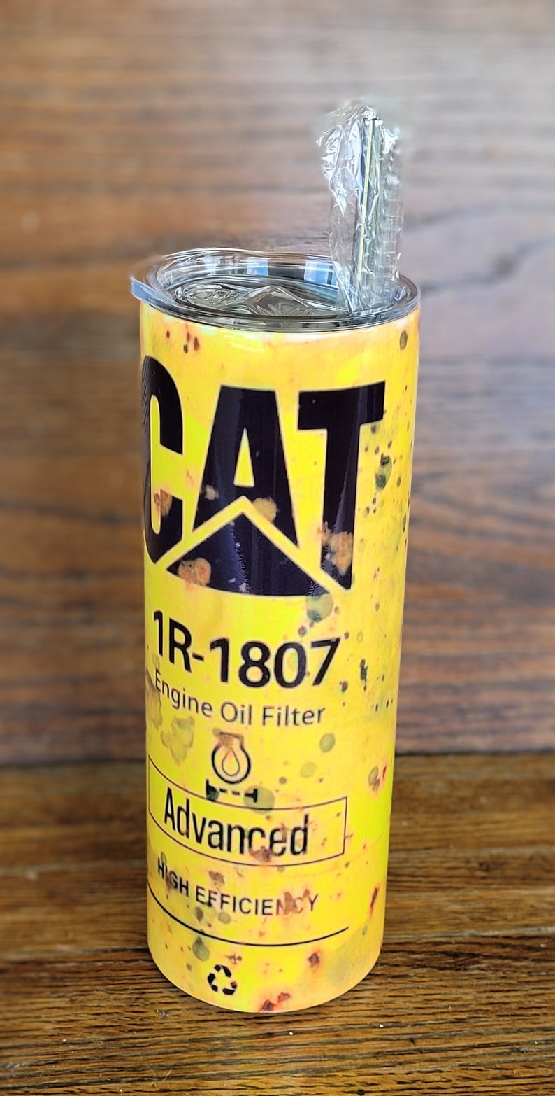 Może przedstawiać: Ż&oacute;łty kubek z logo CAT i napisem "1R-1807 Engine Oil Filter Advanced High Efficiency". Kubek ma przezroczystą pokrywkę i słomkę. Projekt ma postarzany, zużyty wygląd.