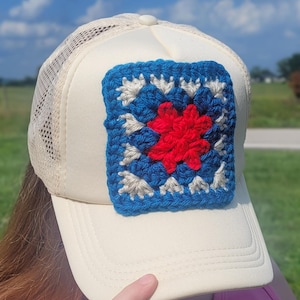 May include: A cream-colored trucker hat featuring a square, multi-colored crochet patch on the front. The patch displays a vibrant red center surrounded by layers of dark blue and light cream yarn in a classic granny square pattern.