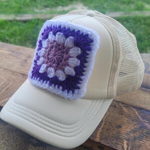 May include: A cream-colored trucker hat featuring a square crochet patch on the front panel. The patch displays a multi-colored granny square pattern in shades of white, light purple, and dark purple, stitched onto the center of the cap.