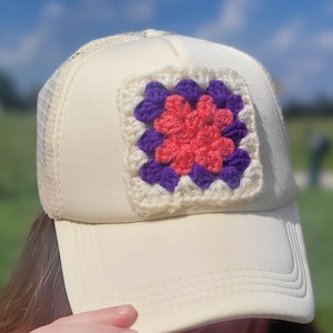 May include: A cream-colored trucker hat featuring a square crochet granny square patch on the front panel. The patch is composed of a coral pink center surrounded by a border of deep purple yarn, all framed by an outer edge of cream-colored yarn.