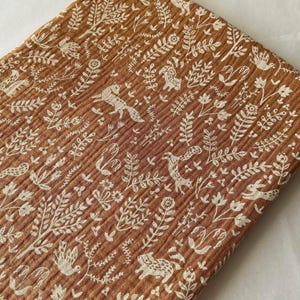 May include: A folded piece of brown fabric with a repeating pattern of white woodland creatures and foliage. The fabric has a textured, ribbed appearance. The design includes foxes, birds, deer, and rabbits among the leaves and flowers.