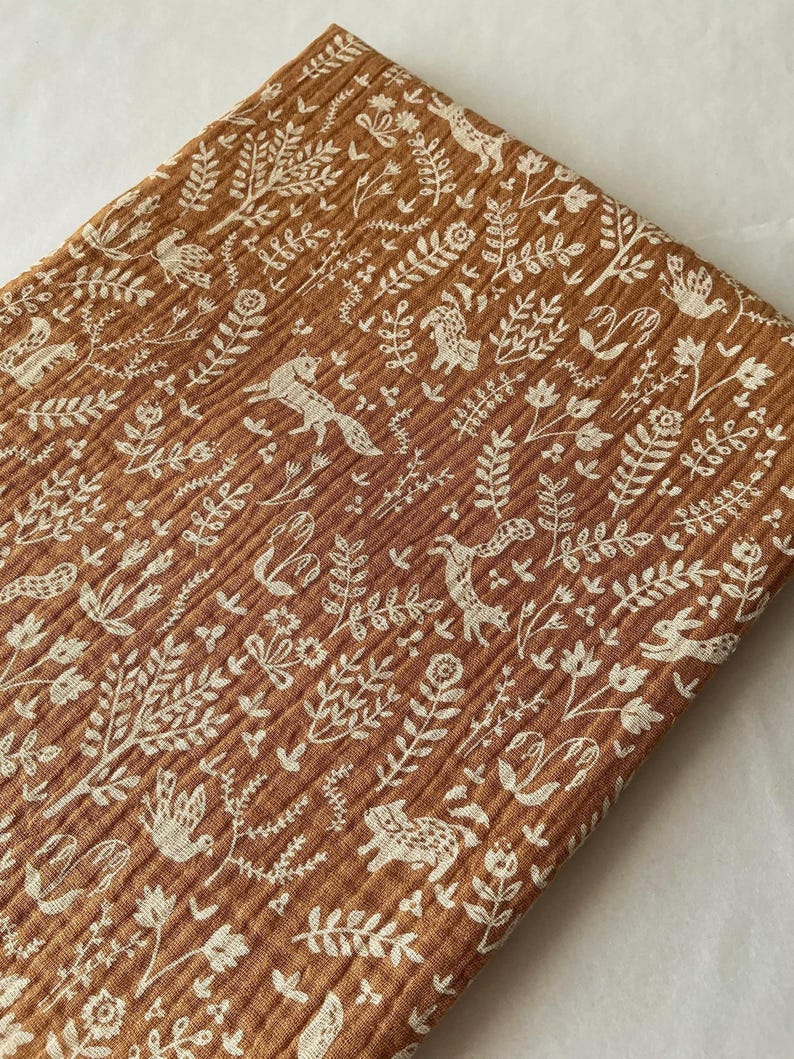 May include: A folded piece of brown fabric with a repeating pattern of white woodland creatures and foliage. The fabric has a textured, ribbed appearance. The design includes foxes, birds, deer, and rabbits among the leaves and flowers.