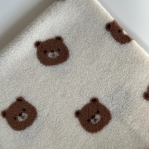 May include: A cream-colored sherpa fleece fabric featuring a repeating pattern of small, brown, fuzzy bear faces. The material has a thick, textured, and plush pile surface with the bear motifs integrated into the fabric design throughout.
