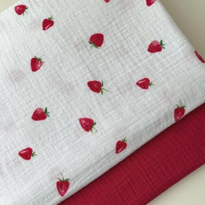 Könnte beinhalten: Zwei Stofflagen, eine weiße und eine rote. Die weiße Lage ist mit roten Erdbeeren und grünen Stielen gemustert. Die rote Lage ist einfarbig, dunkler. Beide Stoffe scheinen weich und strukturiert zu sein.