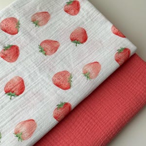 Könnte beinhalten: Zwei Stoffstücke. Die obere Schicht ist weiß mit einem Muster aus roten Erdbeeren mit grünen Blättern. Die untere Schicht ist einfarbig korallenfarben. Die Stoffe scheinen weich und strukturiert zu sein.