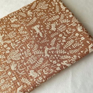 May include: Folded brown fabric with a repeating pattern of white woodland creatures, including foxes, birds, and rabbits, amidst floral and leaf designs. The fabric has a textured, crinkled appearance.