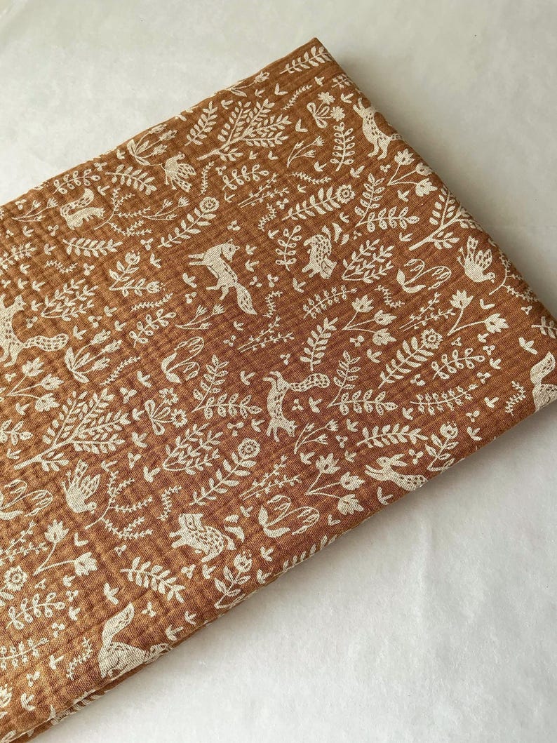 May include: Folded brown fabric with a repeating pattern of white woodland creatures, including foxes, birds, and rabbits, amidst floral and leaf designs. The fabric has a textured, crinkled appearance.