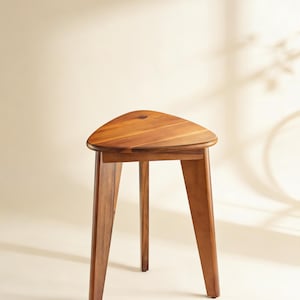 May include: A wooden stool with a triangular seat and three angled legs. The stool is made of a warm-toned wood, and the design is simple and modern. The stool is approximately 18 inches tall.