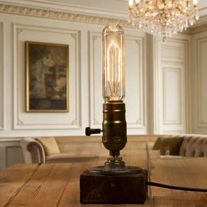 May include: A vintage-style desk lamp with a clear glass bulb and a brass base. The lamp is mounted on a dark wooden block and has a black cord. The bulb is lit, emitting a warm, inviting glow. The lamp is on a wooden table.