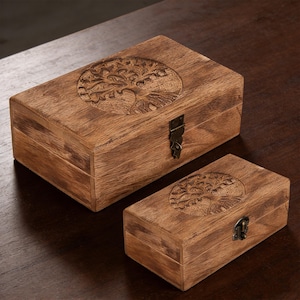 Puede incluir: Dos cajas de madera con un diseño de árbol de la vida tallado en las tapas. Las cajas son rectangulares con una tapa con bisagras y un cierre de metal. La madera tiene un tono marrón cálido.
