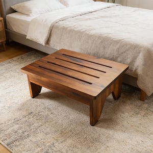 May include: A rectangular, wooden step stool with a rich brown finish. The stool has four legs and a top with four evenly spaced slots. The stool is placed on a patterned rug in front of a bed.