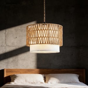 May include: A hanging pendant light with a woven, natural fiber shade. The shade has a geometric pattern and a white fabric diffuser. The light hangs from a rope cord, and is above a wooden bed frame with white pillows.