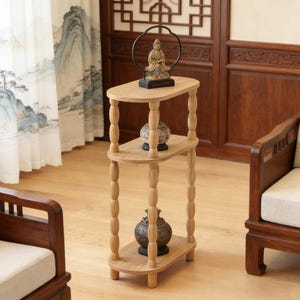 May include: A three-tiered, light-colored wooden side table with an oval top and shelves. The table features turned legs and holds decorative items, including a small statue and two ornate vases. The table is 30 inches tall.