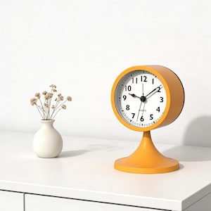 Puede incluir: Un reloj de escritorio analógico amarillo con esfera blanca y números negros. El reloj tiene forma redonda y se apoya sobre una base de pedestal. A la izquierda del reloj hay un pequeño jarrón blanco con flores secas.