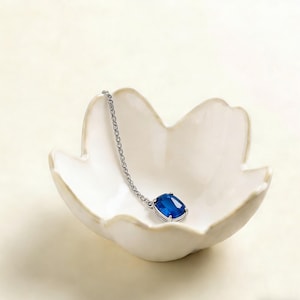May include: A silver necklace with a large, rectangular, deep blue gemstone pendant rests in a white, petal-shaped ceramic dish. The chain is delicate, and the gemstone is set in a silver frame. The dish is on a cream-colored background.