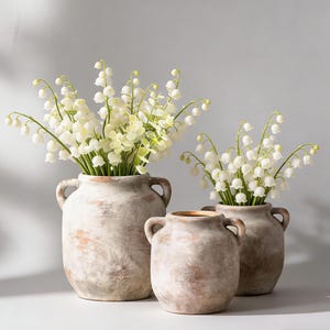 May include: Three rustic, off-white ceramic vases with handles, filled with white lily of the valley flowers. The vases have a distressed, aged appearance. The largest vase is approximately 12 inches tall.