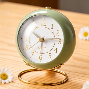 Puede incluir: Un reloj analógico de cuarzo verde salvia con manecillas, números y base dorados. La esfera del reloj es blanca con un borde dorado y la palabra "QUARTZ" impresa en el centro. El reloj es redondo y se apoya en un soporte de metal dorado.