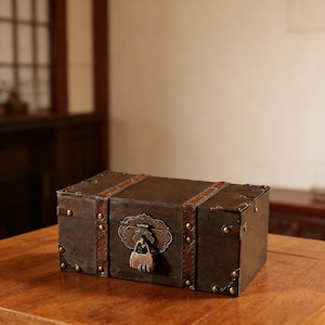 Puede incluir: Un cofre de madera marrón oscuro con correas de cuero y herrajes de latón. El cofre tiene un cierre decorativo y está sobre una superficie de madera. El cofre es rectangular y parece vintage.