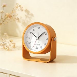 May include: A small, orange quartz analog clock with a white face and black numerals. The clock has a curved base and a silver-colored knob on the side. The clock's hands are black, with a red second hand.