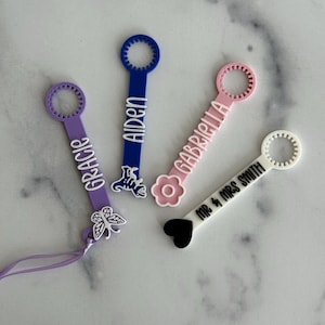 May include: Four colorful bubble wands with personalized names and designs. The wands are purple, blue, pink, and white. The purple wand has a butterfly, the blue wand has a floral design, the pink wand has a flower, and the white wand has a heart.