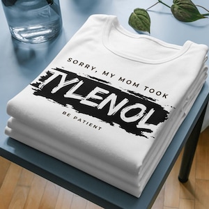 May include: A stack of white t-shirts with the text "SORRY, MY MOM TOOK TYLENOL BE PATIENT" printed in black. The shirts are neatly folded and stacked on a blue surface. The text is in a bold, sans-serif font.