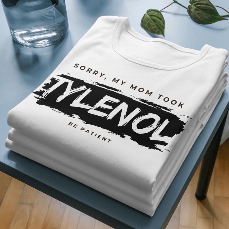May include: A stack of white t-shirts with the text "SORRY, MY MOM TOOK TYLENOL BE PATIENT" printed in black. The shirts are neatly folded and stacked on a blue surface. The text is in a bold, sans-serif font.