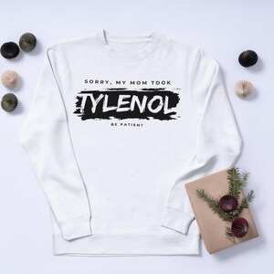 May include: White sweatshirt with the text "SORRY, MY MOM TOOK TYLENOL BE PATIENT" in black. The text is on a black brushstroke background. Decorative items are placed around the sweatshirt.