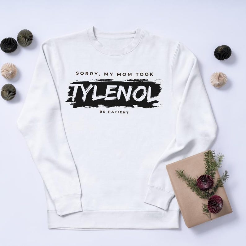 May include: White sweatshirt with the text "SORRY, MY MOM TOOK TYLENOL BE PATIENT" in black. The text is on a black brushstroke background. Decorative items are placed around the sweatshirt.