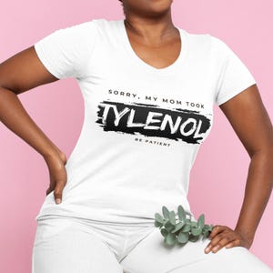 May include: White scoop-neck t-shirt with the text "Sorry, My Mom Took Tylenol, Be Patient" in black. The text is printed over a black brushstroke design. The shirt is worn by a person.