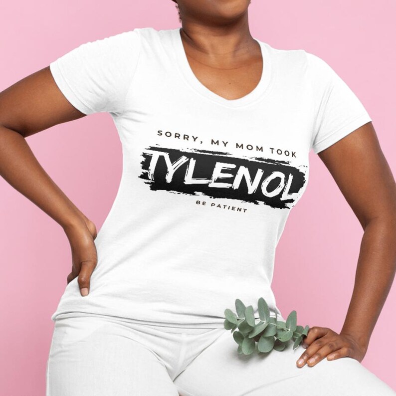 May include: White scoop-neck t-shirt with the text "Sorry, My Mom Took Tylenol, Be Patient" in black. The text is printed over a black brushstroke design. The shirt is worn by a person.