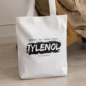 May include: A white canvas tote bag with black text that reads "SORRY, MY MOM TOOK TYLENOL BE PATIENT." The text "TYLENOL" is in a bold, stylized font, and the bag has two white handles.