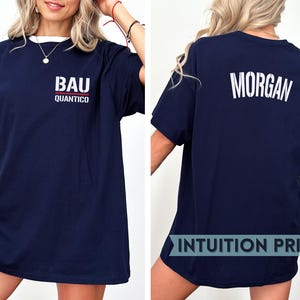 Op de afbeelding: Marineblauw T-shirt met witte tekst "BAU QUANTICO" op de voorkant. De achterkant van het shirt toont de naam "MORGAN" in het wit. Het shirt heeft een witte kraag en is een casual stijl.